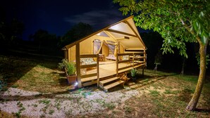 Una tienda acogedora y bien iluminada con terraza de madera, un sofá y una mesa de comedor, rodeada de árboles y un cielo nocturno despejado.