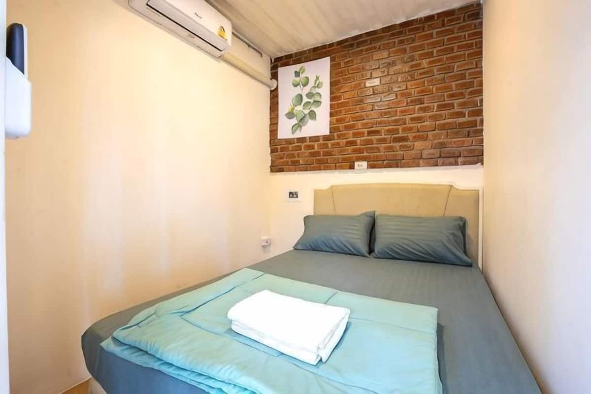 A small, single-bed room with a brick wall, a headboard, and a folded white towel on the bed.