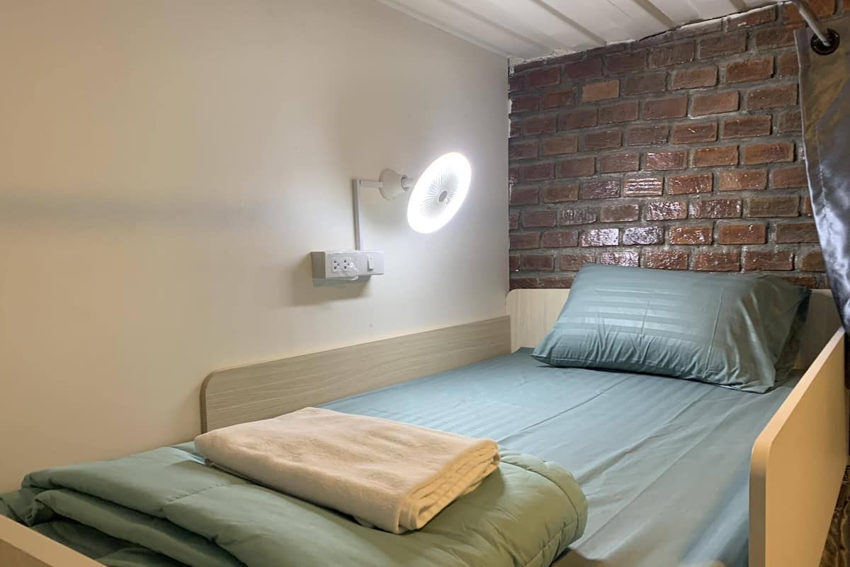 A single bed with a wooden headboard, a green blanket, and a beige towel. There is a wall-mounted light fixture and a brick wall in the background.