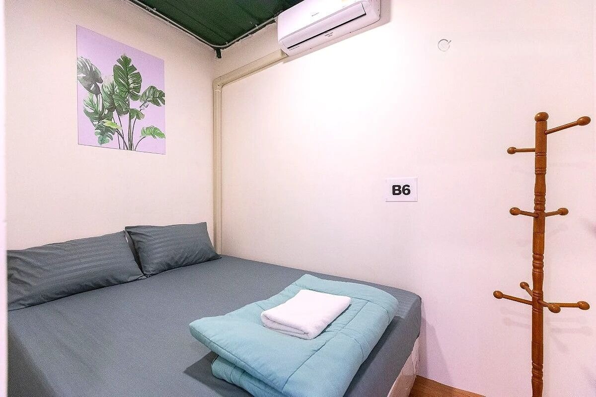 A room with a bed, a wall-mounted air conditioner, a coat rack, and a room number sign.