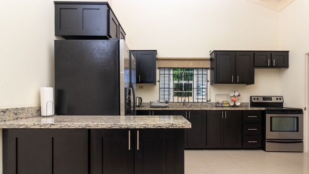 A modern kitchen with dark cabinetry, a granite countertop, a stainless steel refrigerator, a built-in oven, and a window with blinds. - Lot 40 Draxhall  (St. Ann's Bay)