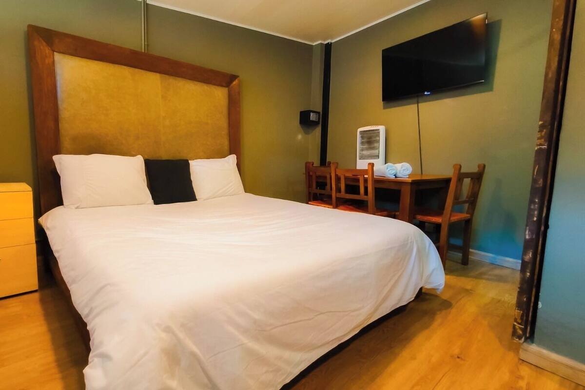 A neatly made bed with white linens, a wooden headboard, a small bedside table with drawers, a chair, a desk with a chair, and a flat-screen TV mounted on the wall.