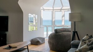 A modern living room with a large bay window overlooking the sea, a comfortable sofa, a small table, and a lamp. - Spectacular Sea View Penthouse Apartment + Parking (Newquay)