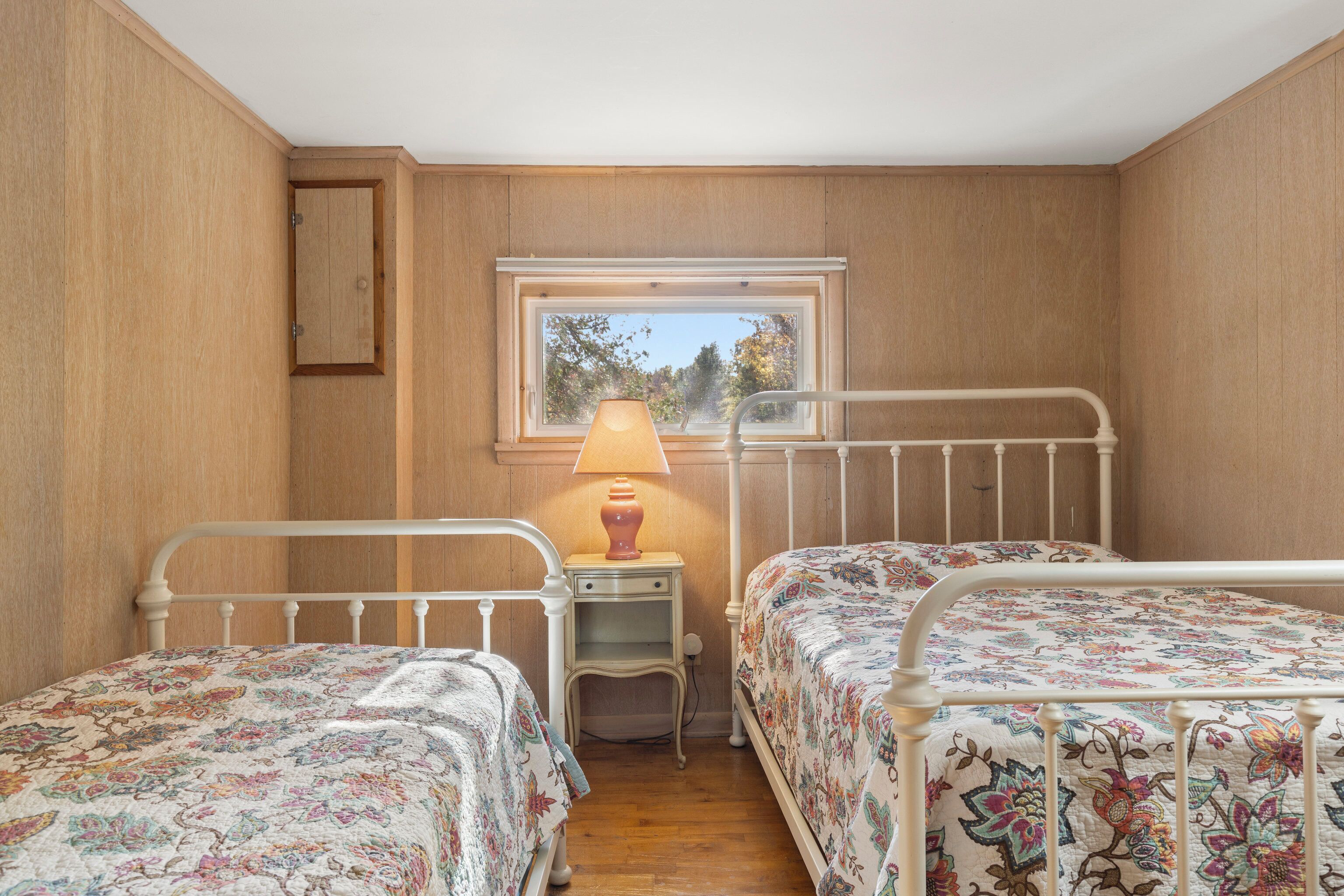 A bedroom with two single beds, a nightstand with a lamp, and a window with a view of trees.