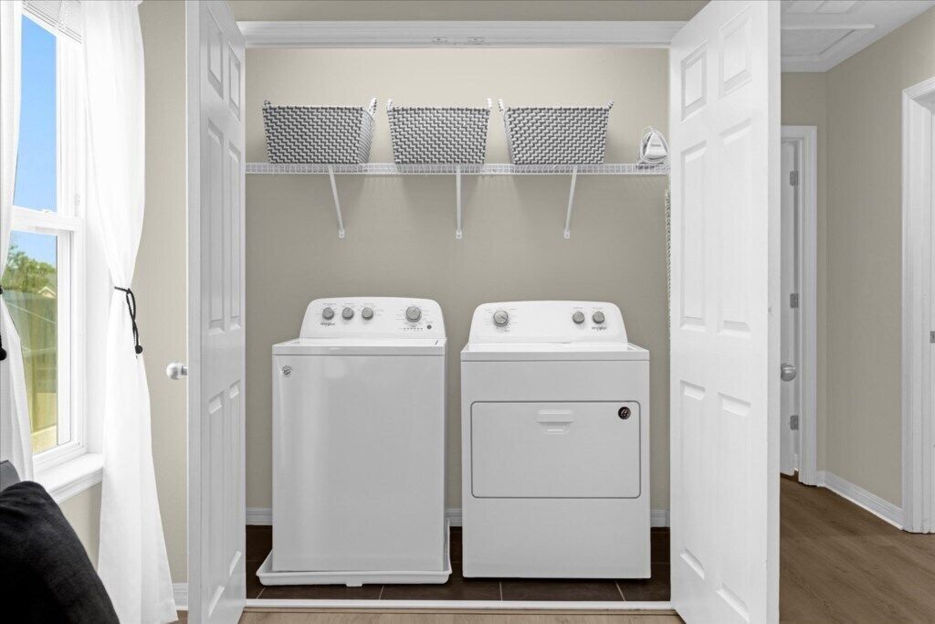Un cuarto de lavandería con dos lavadoras blancas, un estante con ropa colgada y una puerta que da a otra habitación.