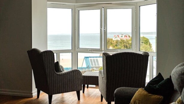 Een kamer met een grote erker met uitzicht op zee, twee fauteuils en een klein tafeltje.