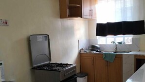 Une cuisine équipée d’une cuisinière à gaz, d’un four et d’un évier. On y trouve une fenêtre avec un rideau noir, des placards en bois et un sol carrelé.