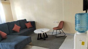 Un salon avec un canapé gris, une table basse ronde, un fauteuil rose et un distributeur d’eau.