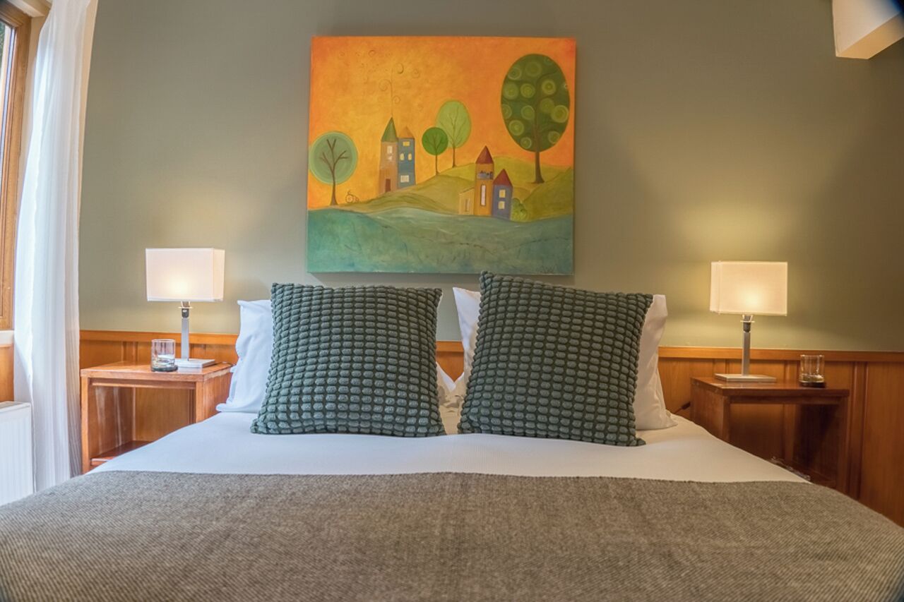 Una habitación de hotel con una cama grande, dos lámparas de noche, un cuadro de un paisaje y una ventana con cortinas.