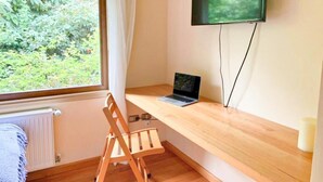 A room with a wooden desk, a laptop, a TV mounted on the wall, a window with a view of greenery, and a chair. - Amadeus Hotel (Frutillar)