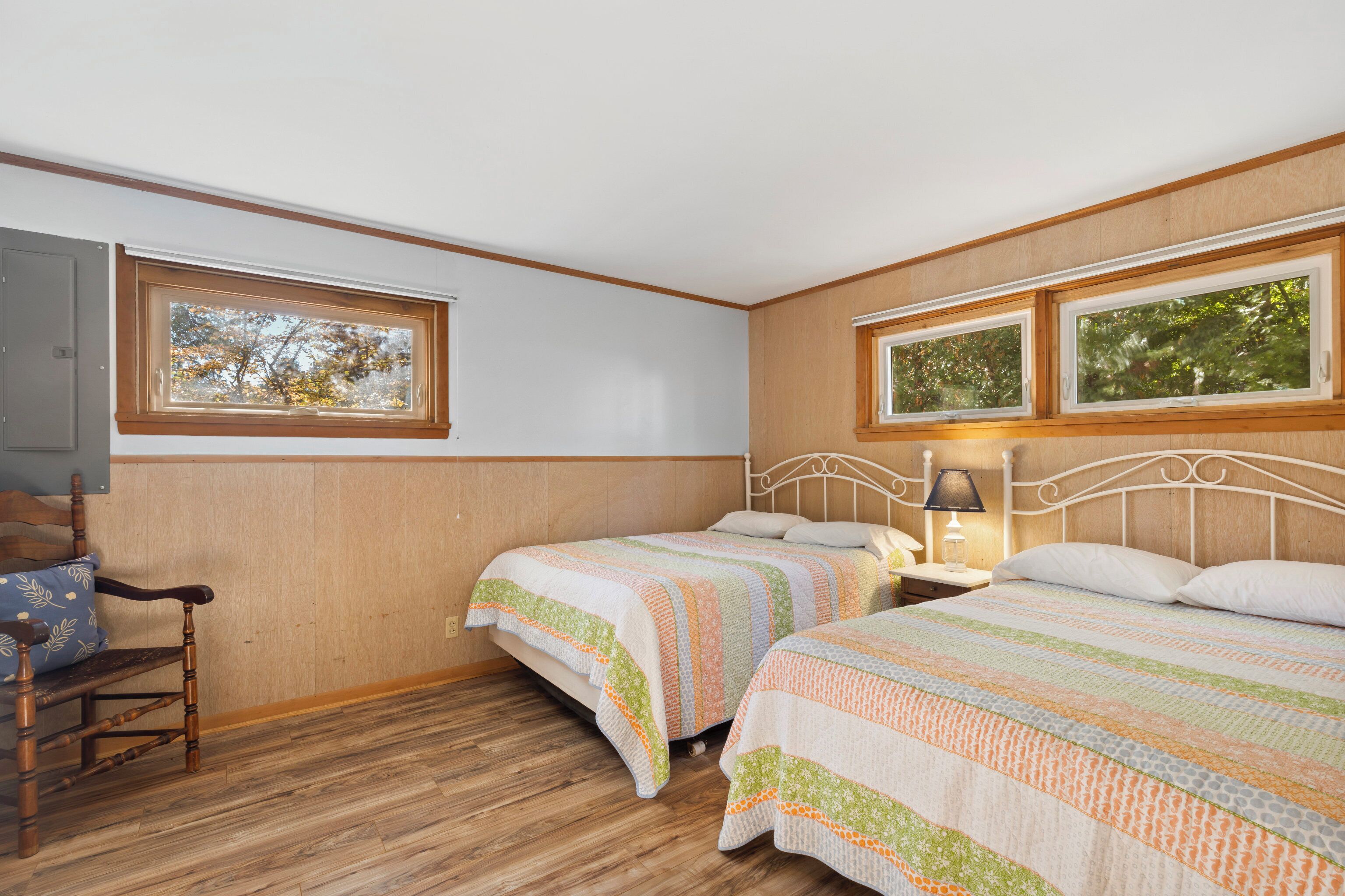 Ein Schlafzimmer mit zwei Betten, Holzboden, einem Sessel und einem Fenster mit Blick auf Bäume.