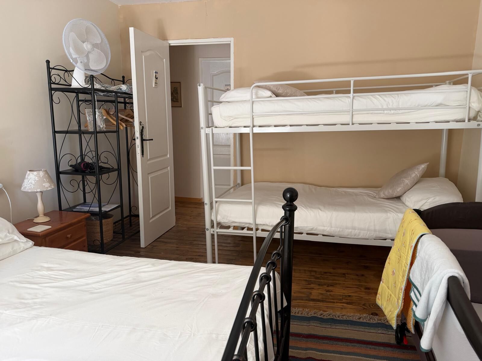 A room with bunk beds, a bedside table with a lamp, a fan, and a door leading to another room.