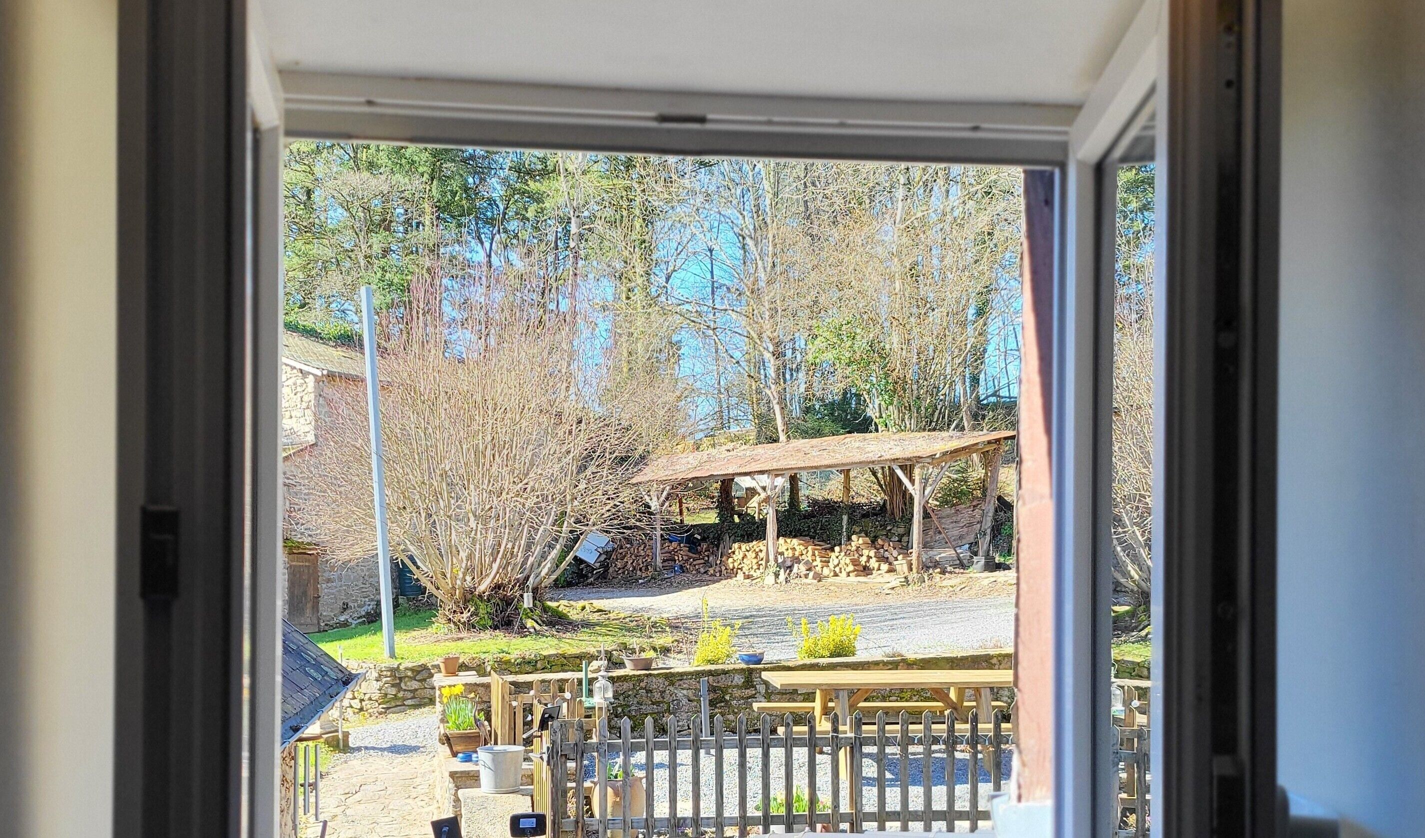 A view through a window of a patio area with a wooden table, chairs, and a stone wall.