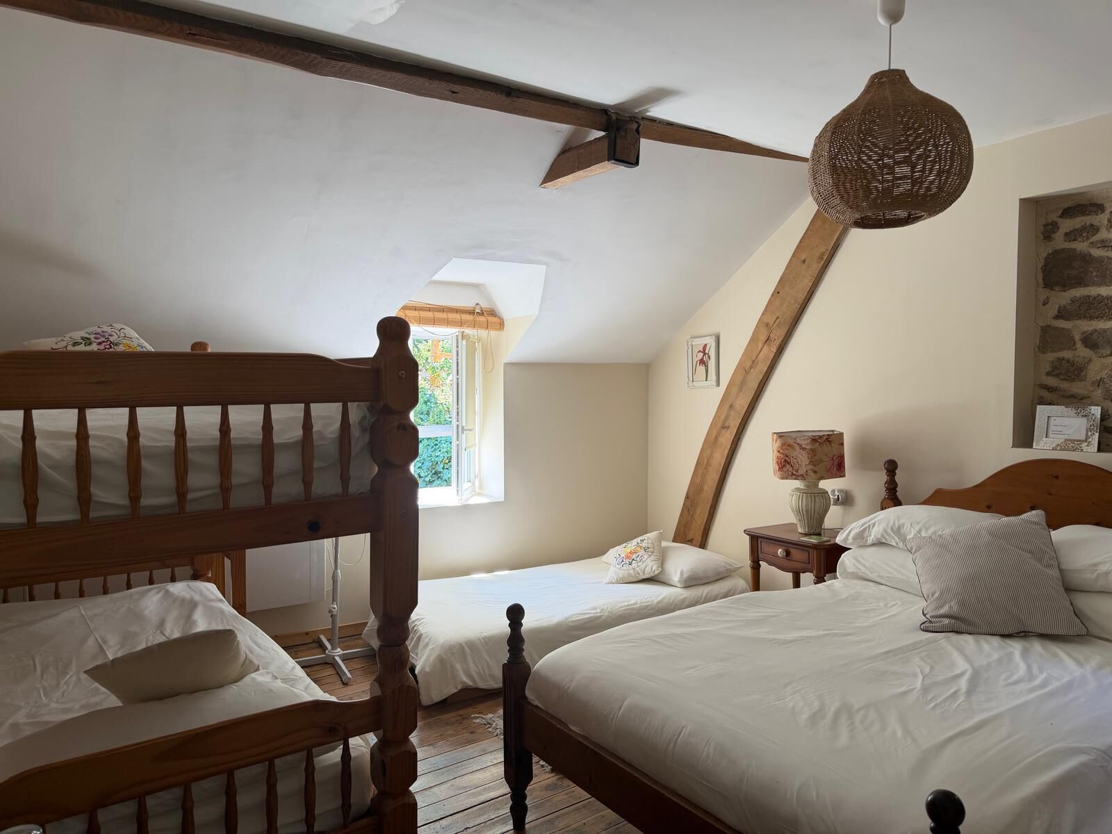 A bedroom with two beds, a wooden headboard, a hanging light fixture, and a window with a view of greenery.