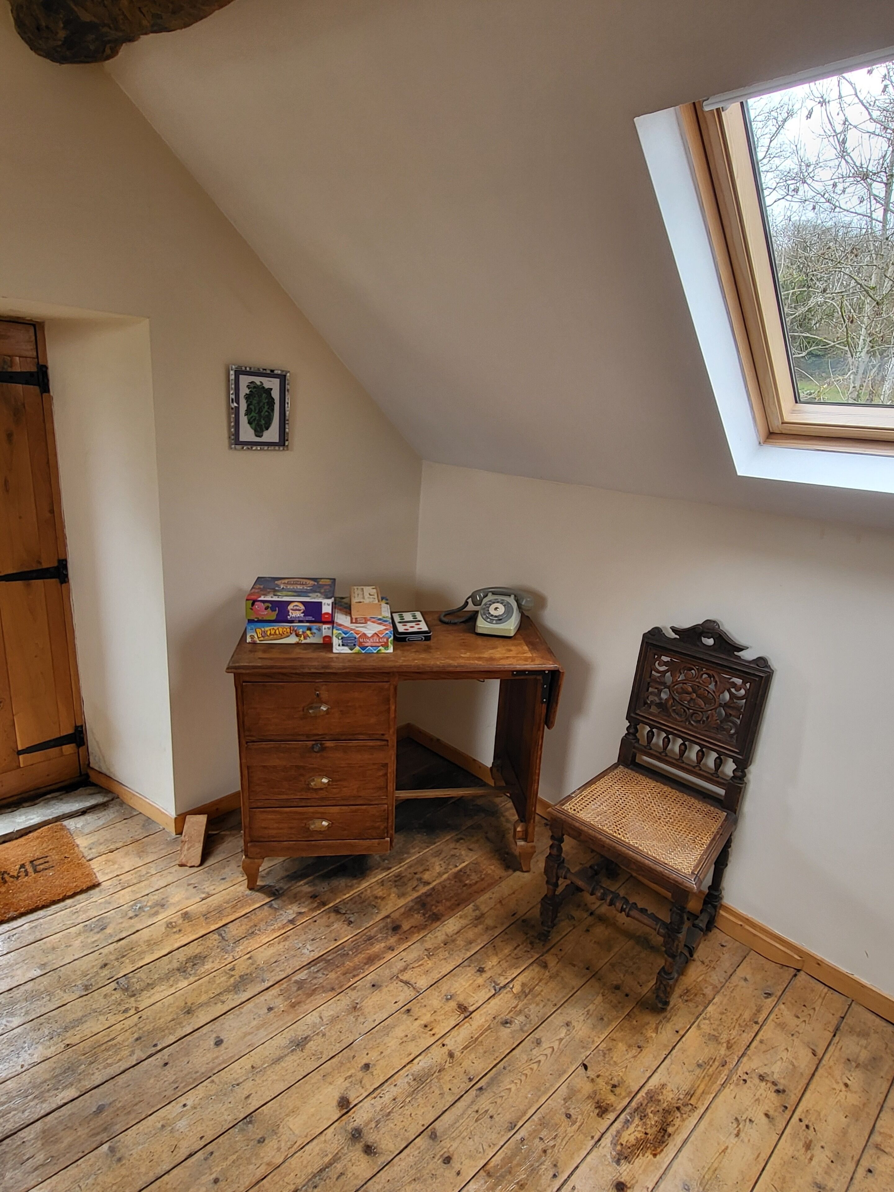 Una habitación con techo inclinado, un escritorio de madera, una silla antigua y una ventana con vistas a árboles.