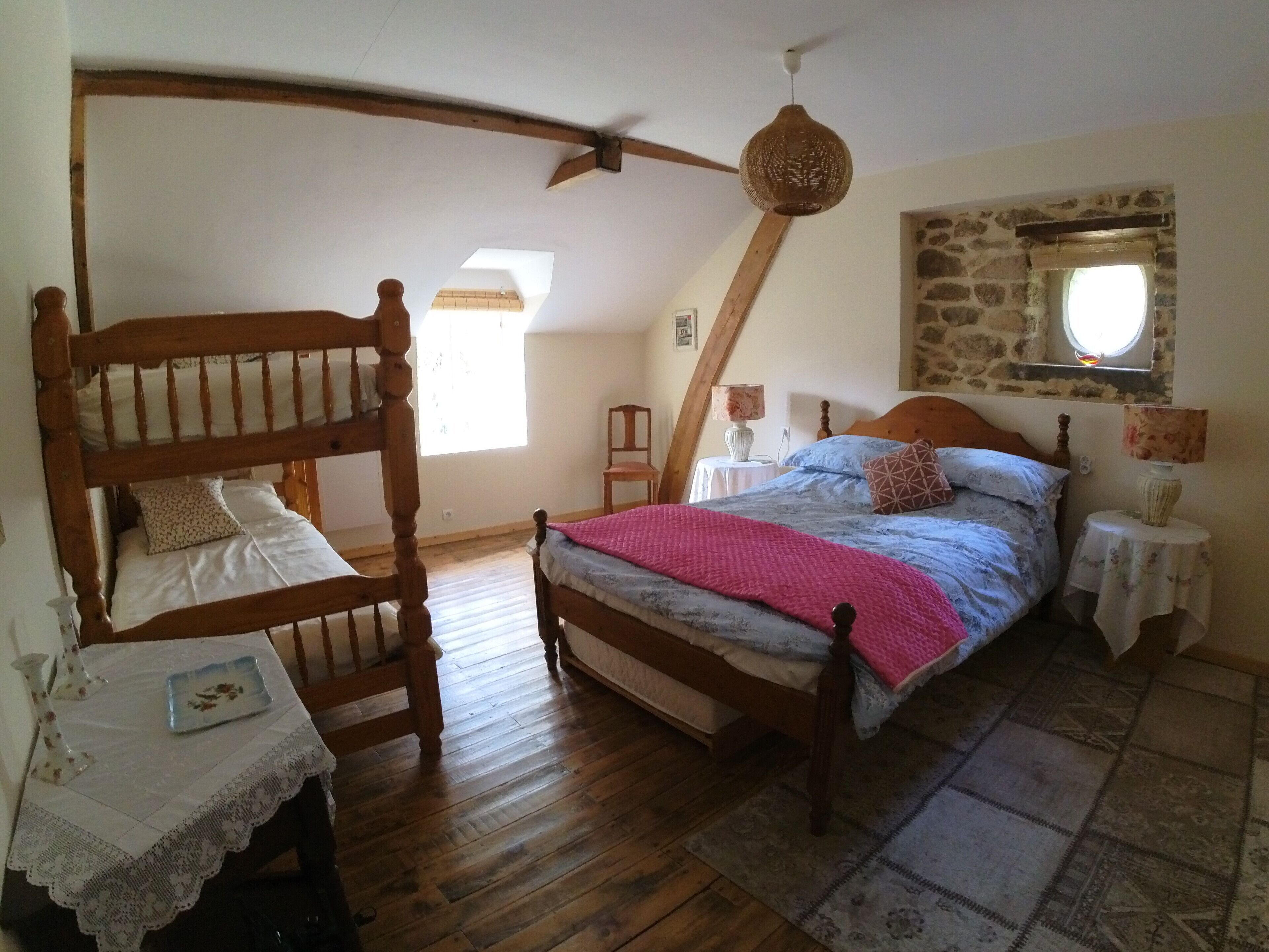 Un dormitorio con una cama litera de madera, una cama individual con una manta rosa, una mesita con un florero y una pared de piedra con una ventana.