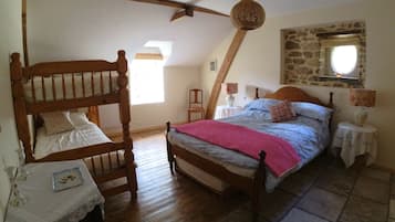 A bedroom with a wooden bunk bed, a single bed with a pink blanket, a small table with a vase, and a stone wall with a window.