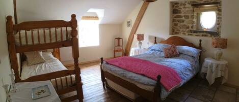 Un dormitorio con una cama litera de madera, una cama individual con una manta rosa, una mesita con un florero y una pared de piedra con una ventana.