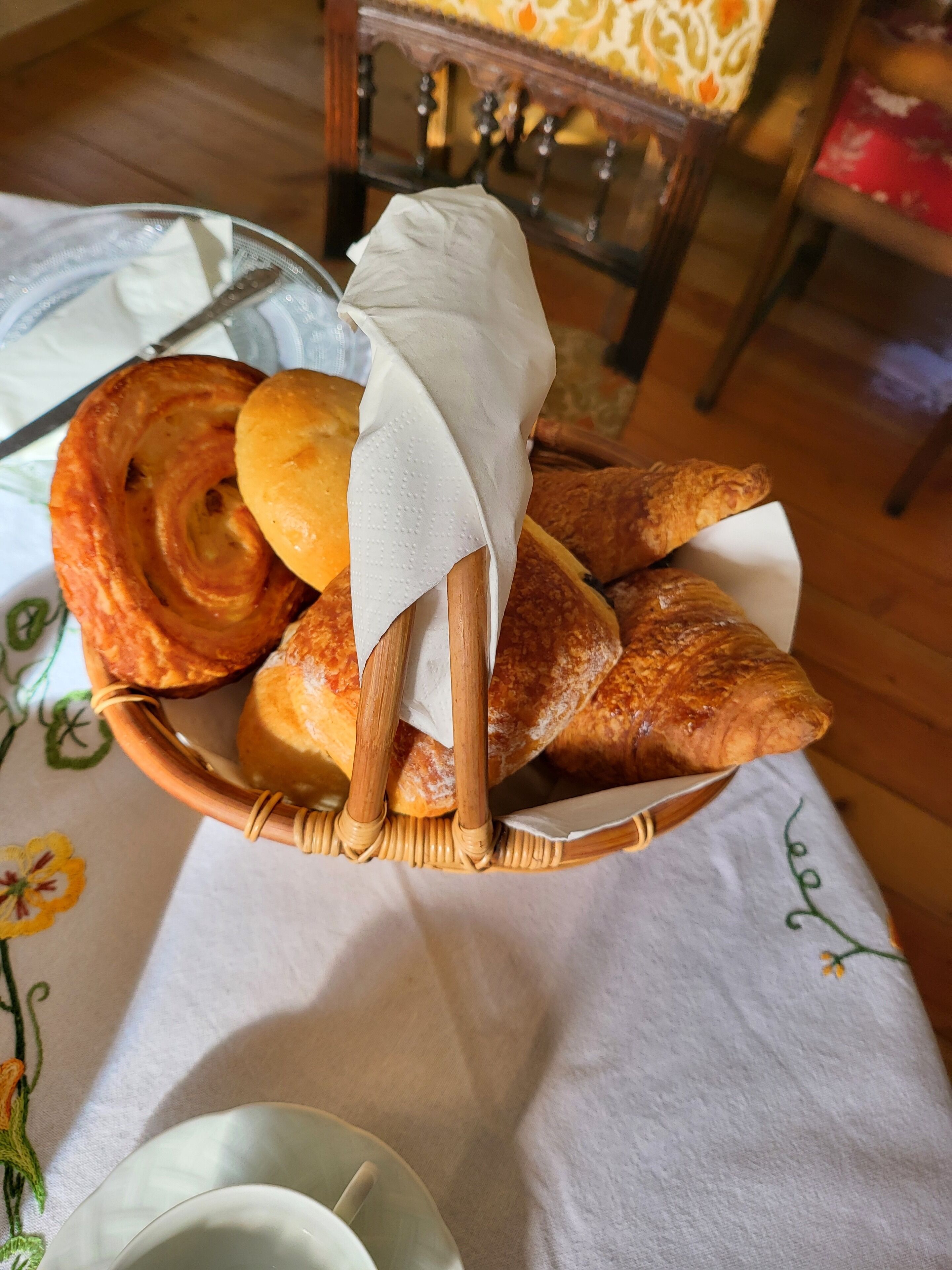 Una cesta de mimbre llena de varios pasteles, incluyendo croissants y un pastel enrollado, sobre una mesa con mantel floreado.