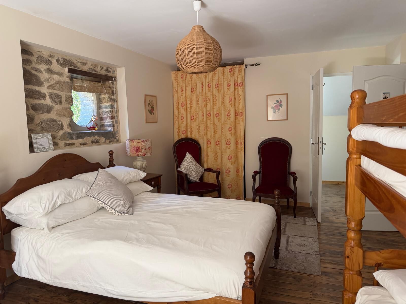 A bedroom with a large bed, two chairs, a hanging lamp, a stone wall, and a door.