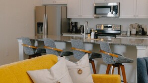 A modern kitchen with white cabinets, a stainless steel refrigerator, a microwave, and a coffee maker. There are four bar stools with grey upholstery and wooden legs. A yellow sofa with a white pillow is in the foreground.