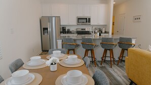 A modern kitchen with white cabinets, a stainless steel refrigerator, and a microwave. There is a dining area with a wooden table set for four, surrounded by grey upholstered chairs.