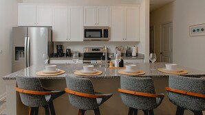 A modern kitchen with white cabinets, stainless steel appliances, and a granite countertop. There are four bar stools with woven seat pads and wooden armrests.