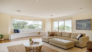 A spacious living room with a large beige sectional sofa, a round coffee table, and a built-in bench with cushions. - Anchors at Mollymook Beach (Mollymook)