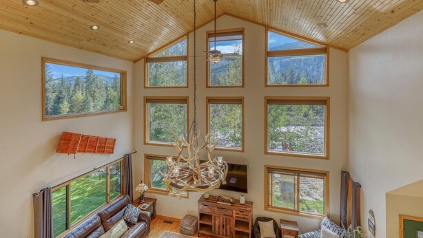 A spacious living room with high ceilings, large windows offering views of a forested landscape, a wooden ceiling, and a chandelier.