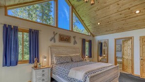 A bedroom with a large bed, wooden ceiling, and a view of trees through the windows.