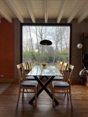 A dining area with a glass table and wooden chairs, a hanging light fixture, and a view of trees through a large window. - Vrbo Property (Lévignacq)