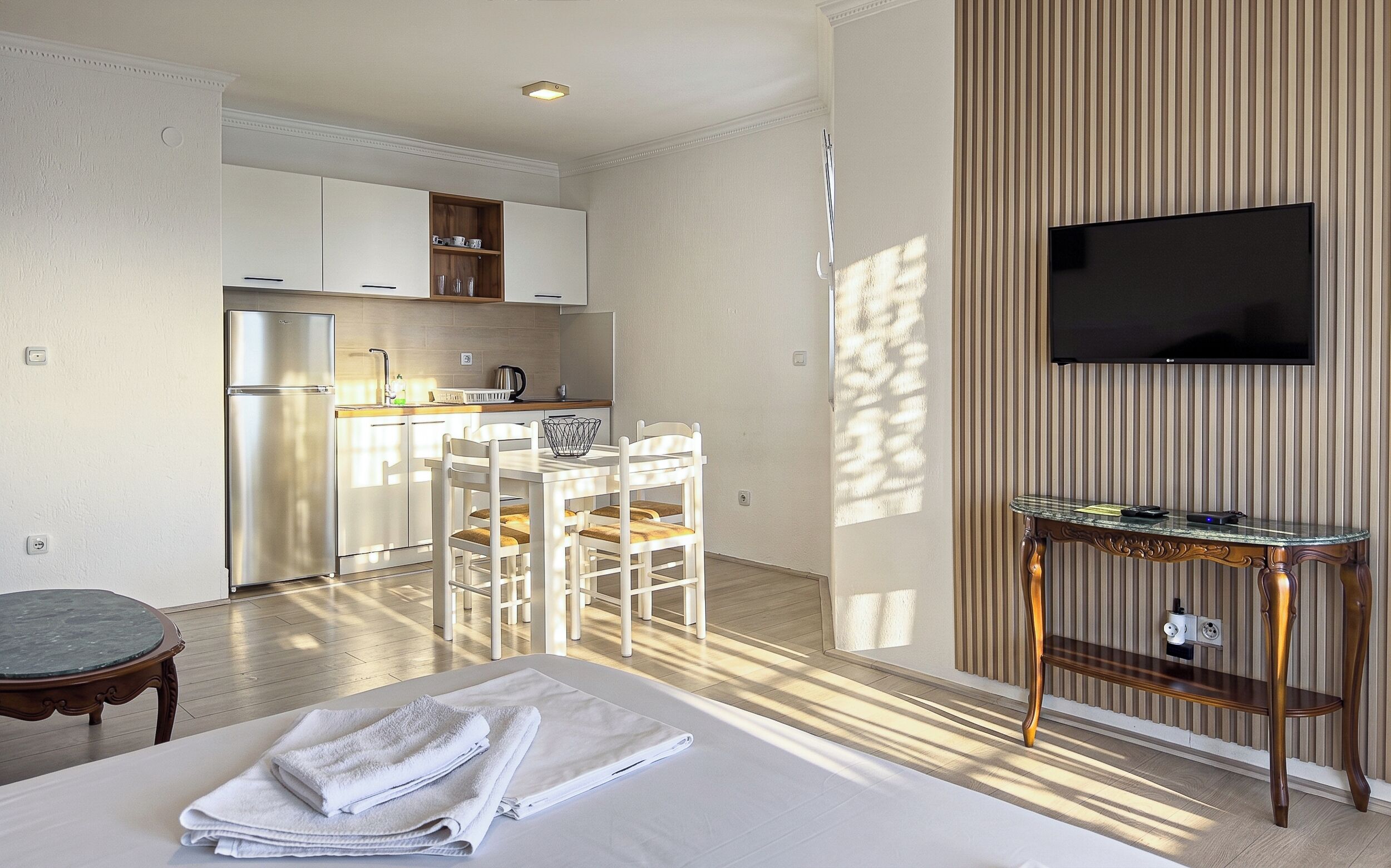 A hotel room with a bed, a small table, a TV, a kitchen area, and a dining table.