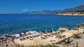 Une station balnéaire avec des transats et des parasols sur une plage rocheuse, offrant une vue sur la mer et les montagnes.