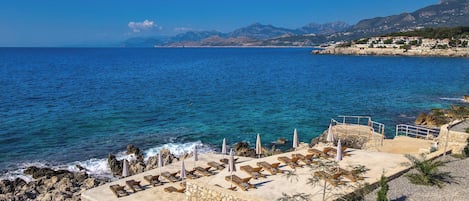 A coastal resort with sun loungers and umbrellas on a rocky shore, overlooking the sea and mountains.