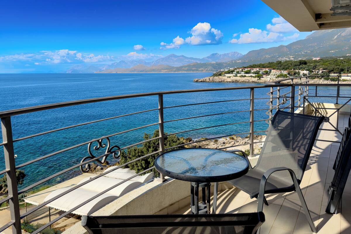 A balcony with a table and chairs overlooking a coastal view with mountains in the distance.