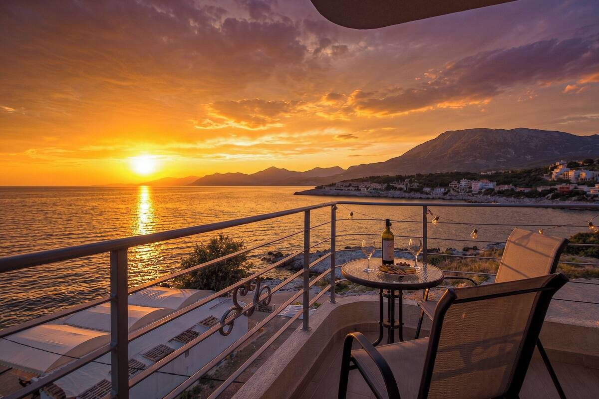 A balcony with a table, chairs, and a bottle of wine, overlooking a sunset over the sea with mountains in the background.
