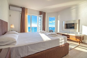 A hotel room with a large bed, a television, a balcony with curtains, and a view of the sea.