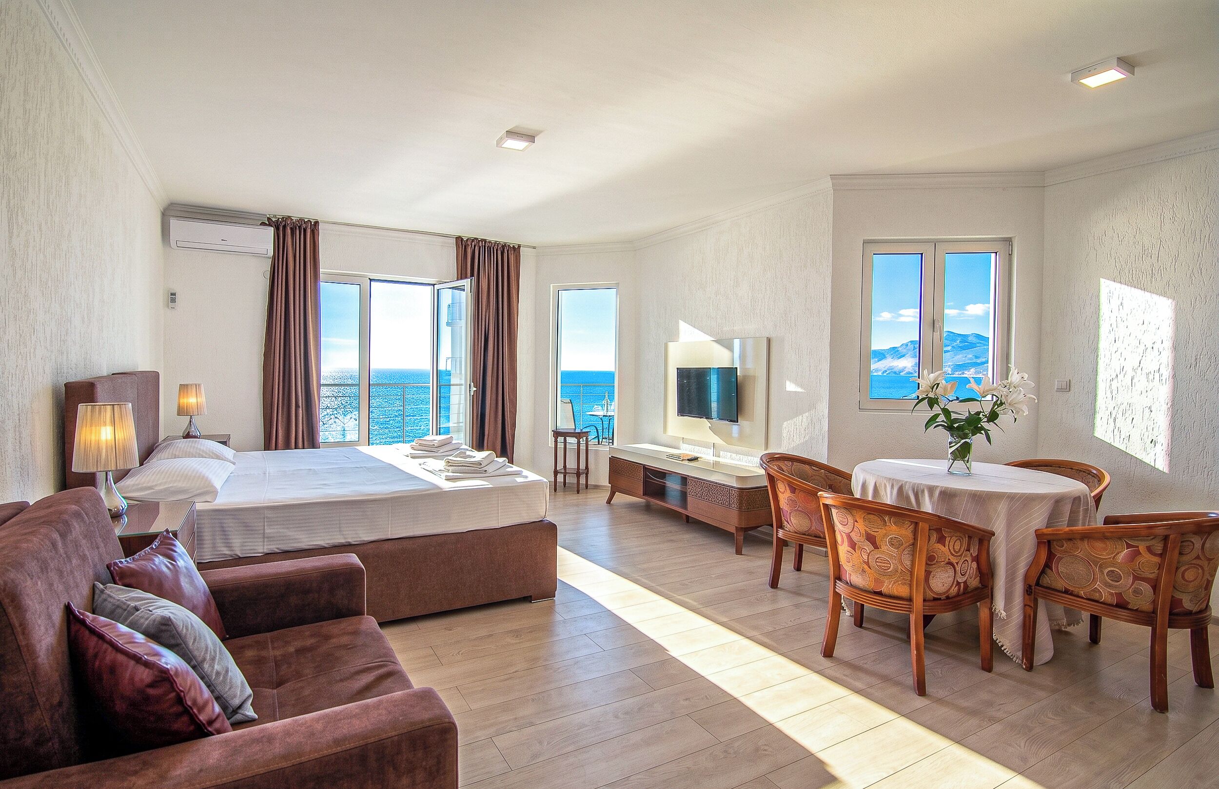 A hotel room with a bed, a sofa, a dining table with chairs, a TV, and a view of the sea.