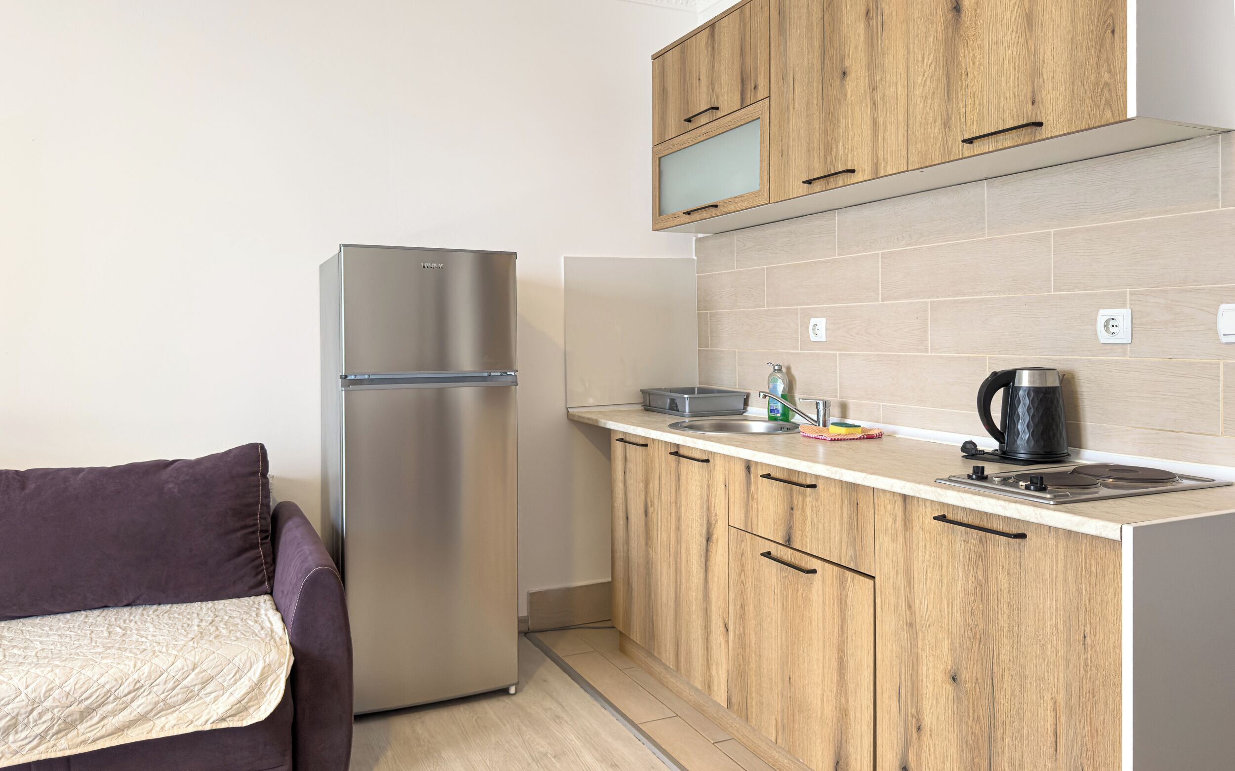 A compact kitchen with wooden cabinets, a stainless steel refrigerator, and a countertop with a kettle and dish soap.