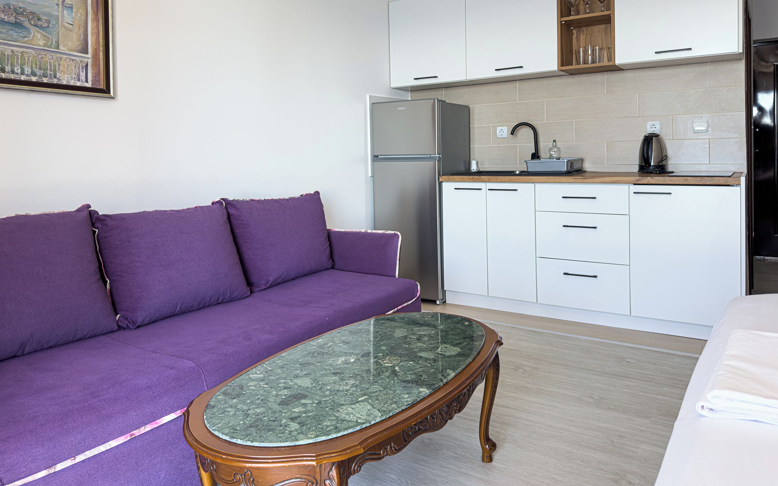 A compact living space with a purple sofa, a marble coffee table, a refrigerator, and a kitchenette with white cabinets and a wooden countertop.