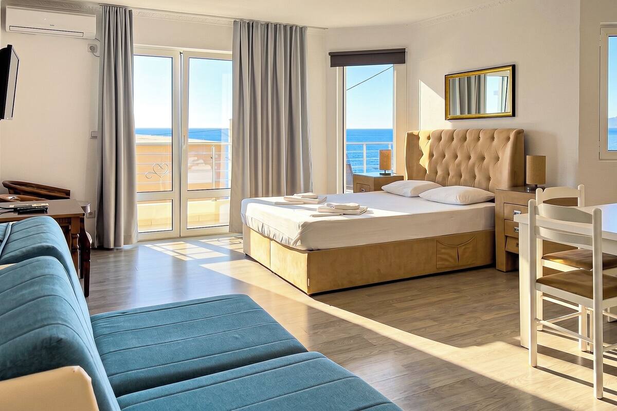 A hotel room with a bed, a sofa, a dining table, and a view of the sea.