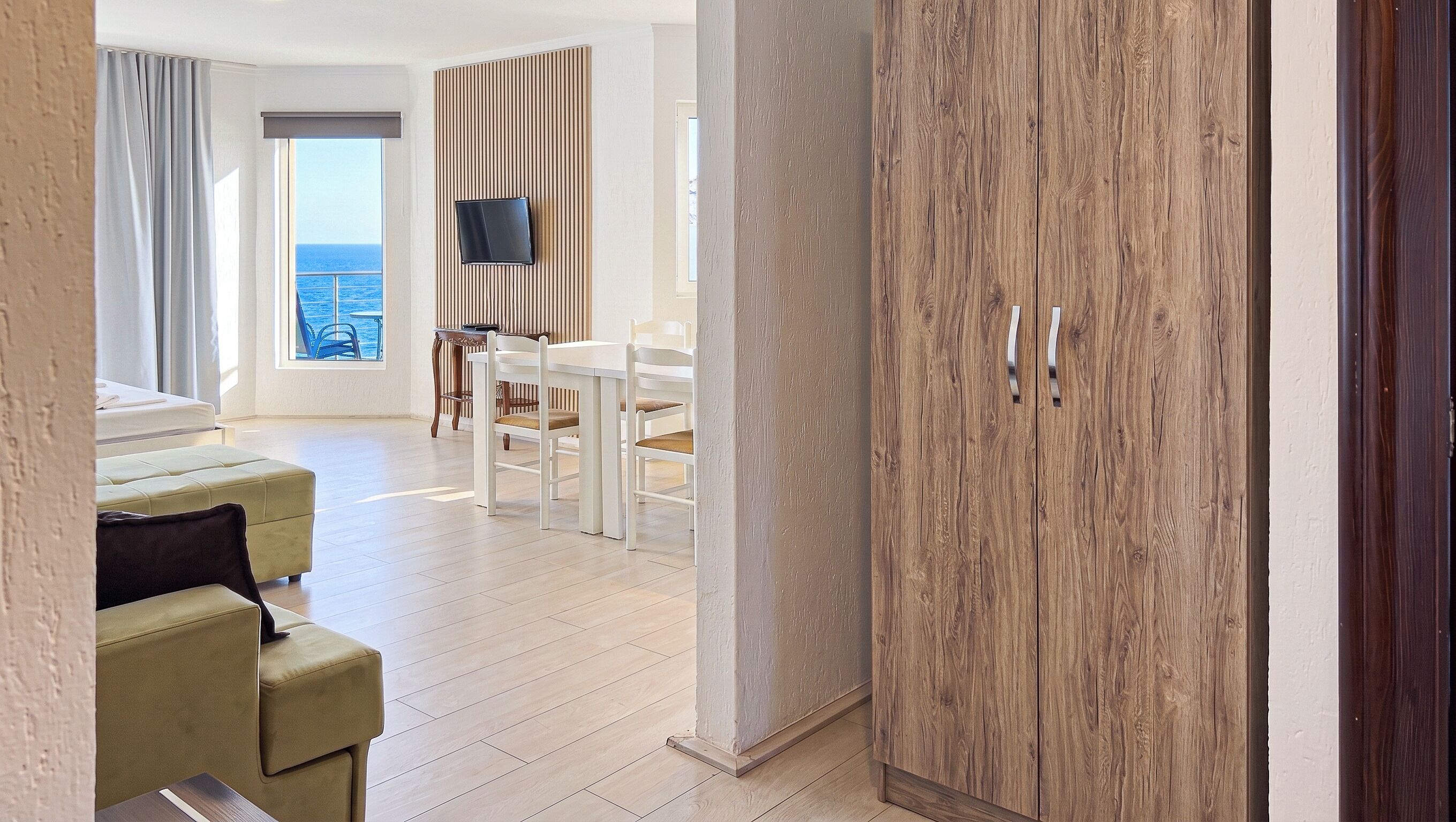 Een moderne hotelkamer met een houten garderobe, een zithoek met een tafel en stoelen, een aan de muur bevestigde televisie, en uitzicht op de zee door het raam.