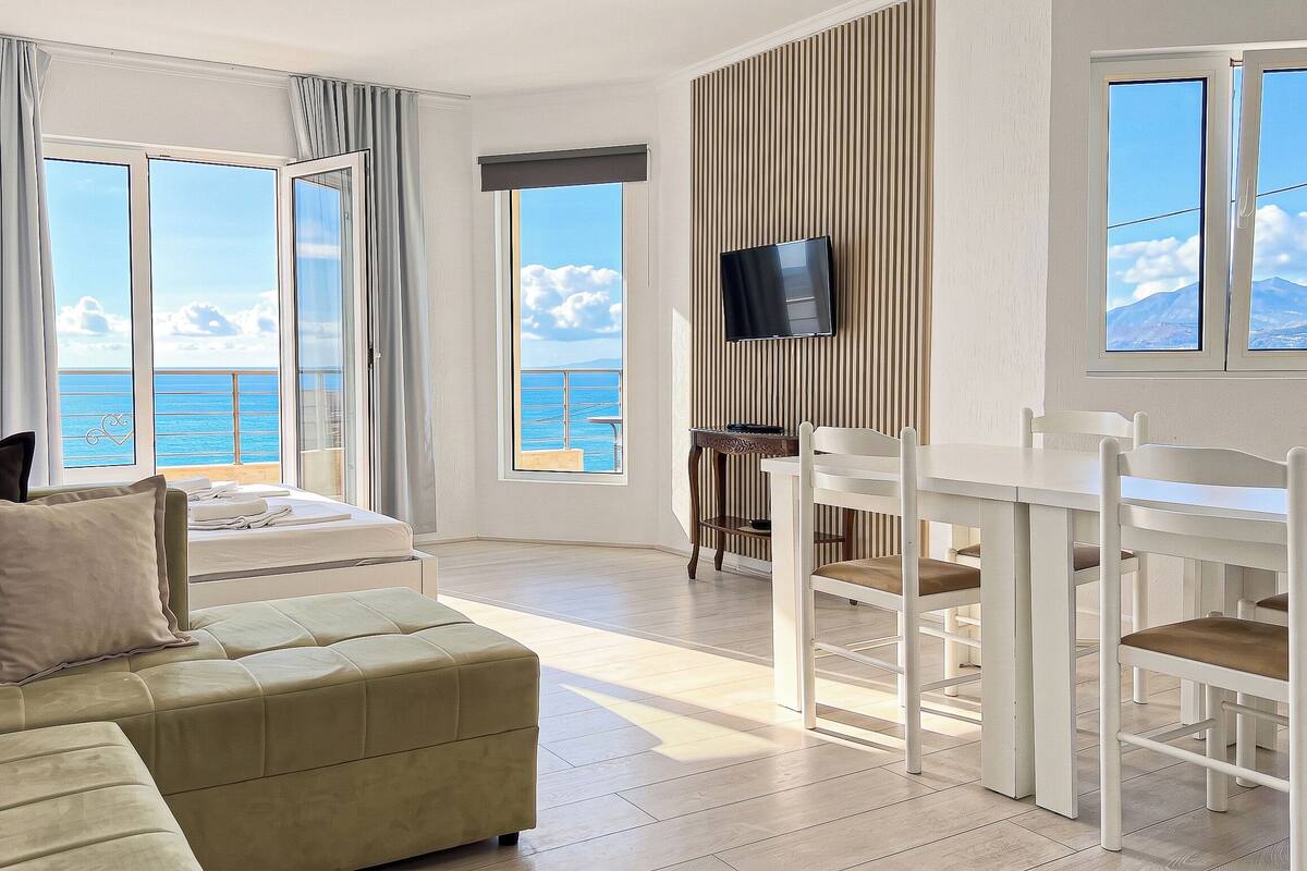 A room with a bed, a sofa, a dining table with chairs, a TV, and large windows with a view of the sea.