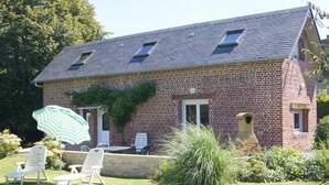 Ein Ziegelhaus mit geneigtem Dach, zwei Fenstern und einer Tür. Vor der Haustür befindet sich eine Terrasse mit zwei weißen Lounge-Stühlen und einem gestreiften Sonnenschirm.