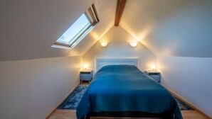 Ein Schlafzimmer mit Schrägdecke, einem Dachfenster, einem Bett mit blauer Bettdecke, zwei Nachttischen mit Lampen und einem Holzboden.