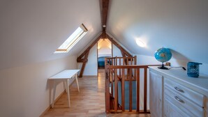 Ein schmales Dachzimmer mit Holzbalken, einem weißen Schreibtisch, einem Globus auf einer Kommode und einem Oberlicht.