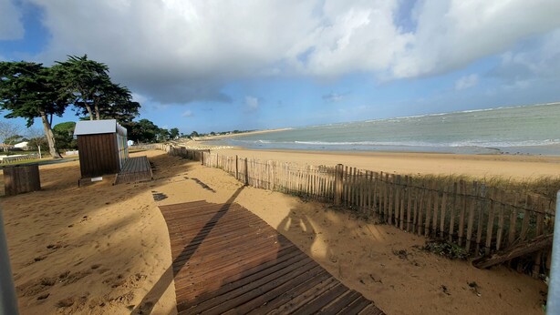 Une promenade en bois mène à une plage de sable, bordée d’une clôture en bois et agrémentée d’une petite construction.