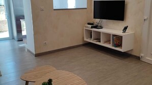 Un salón moderno con un televisor de pantalla plana montado en la pared, un mueble blanco de entretenimiento debajo y dos mesas de madera con una planta en maceta.