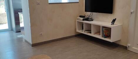 A modern living room with a flat-screen TV mounted on the wall, a white entertainment unit below, and two wooden tables with a potted plant.