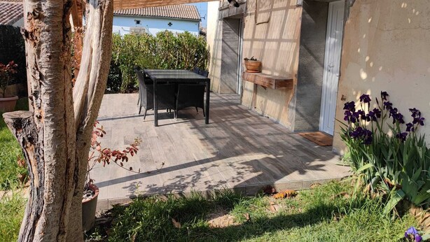 Un patio con una mesa y sillas, una pérgola, un estante con un cuenco y una planta en maceta con flores moradas.